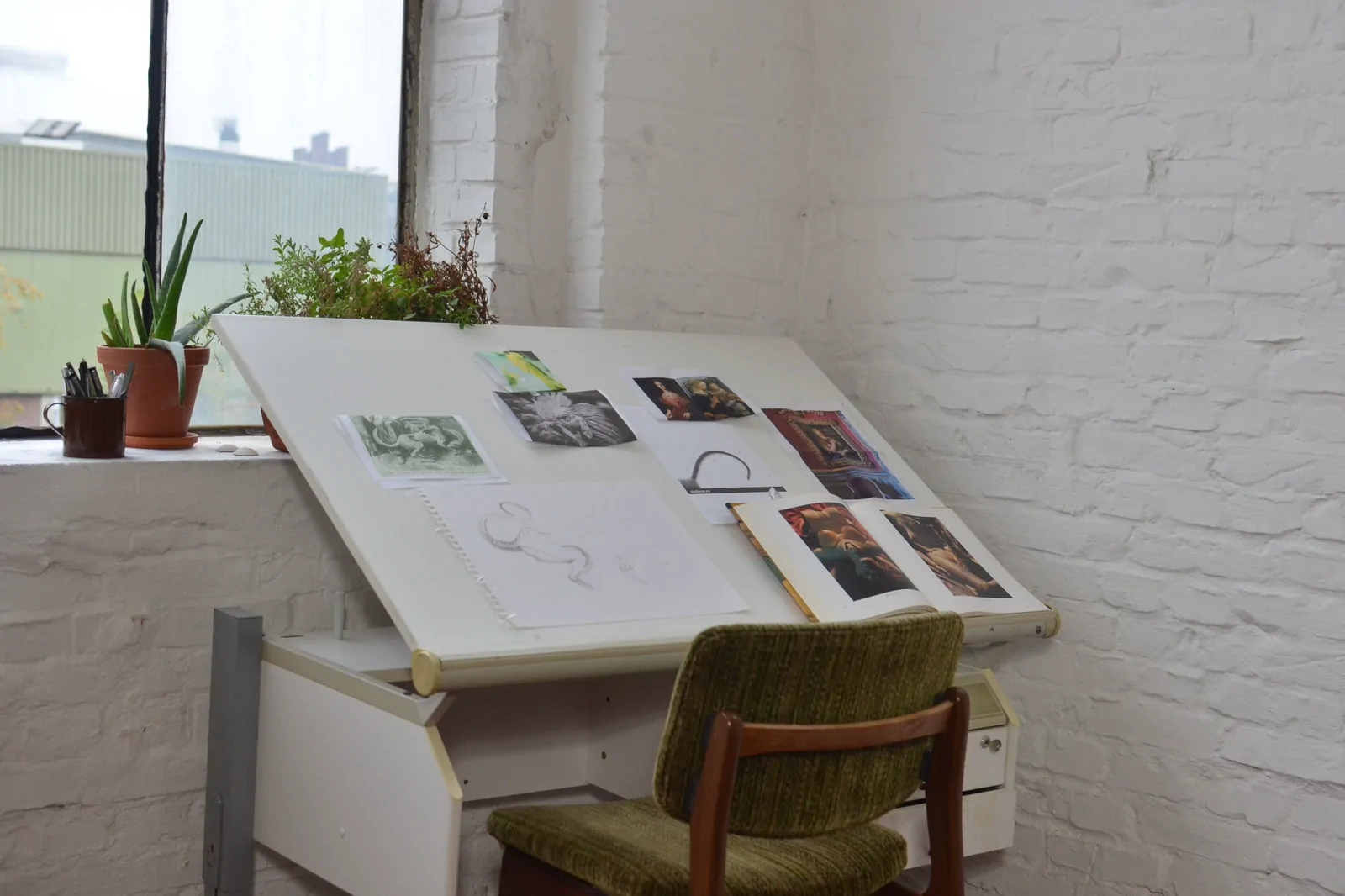 A studio layout table with printed work spread across it, against a white brick wall