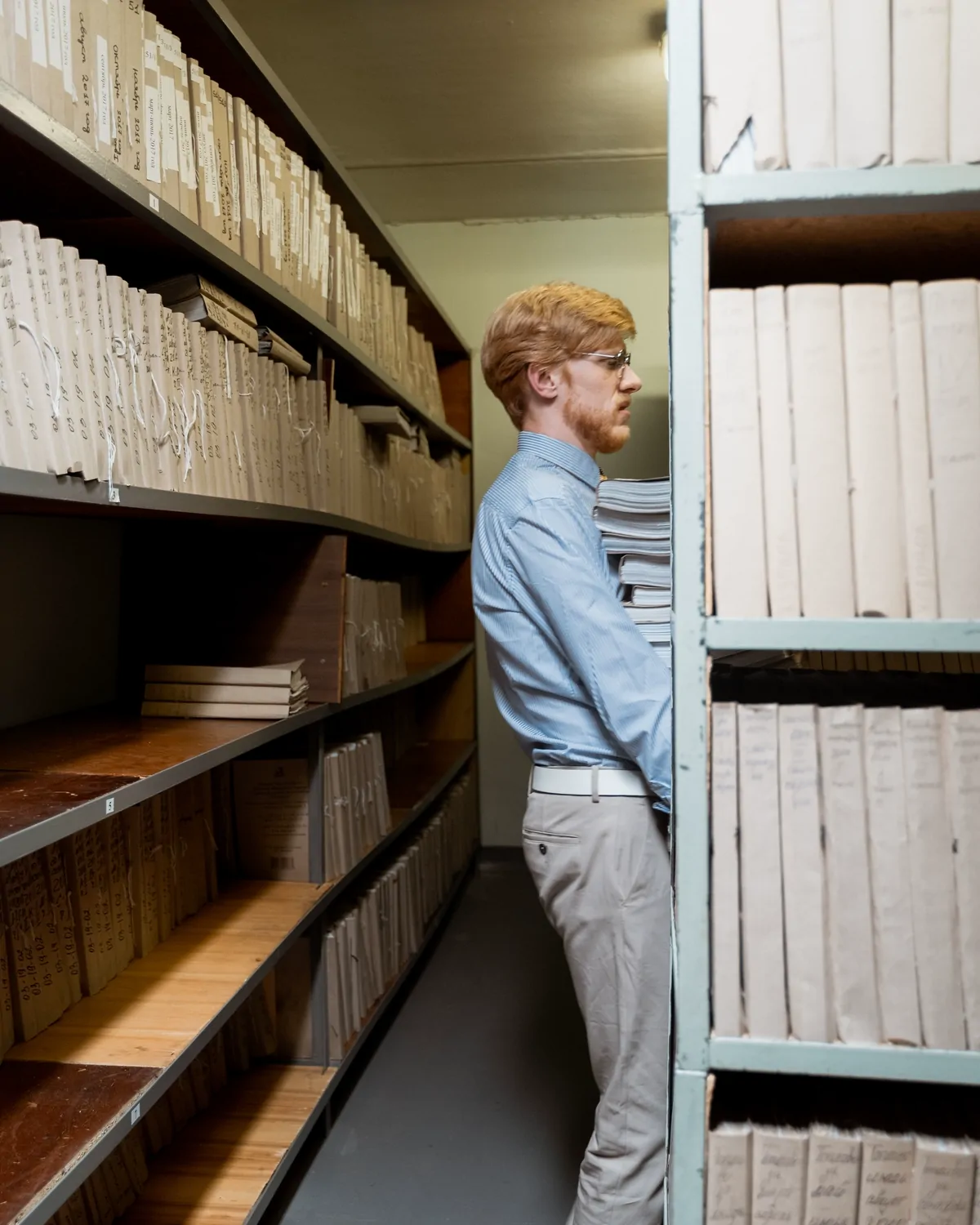 A researcher standing between tall shelves of bound volumes in an archive aisle