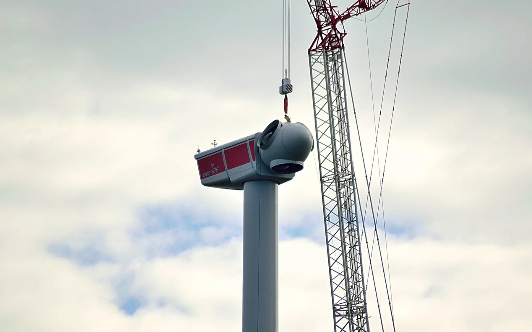 A turbine nacelle suspended from a crane against an overcast sky
