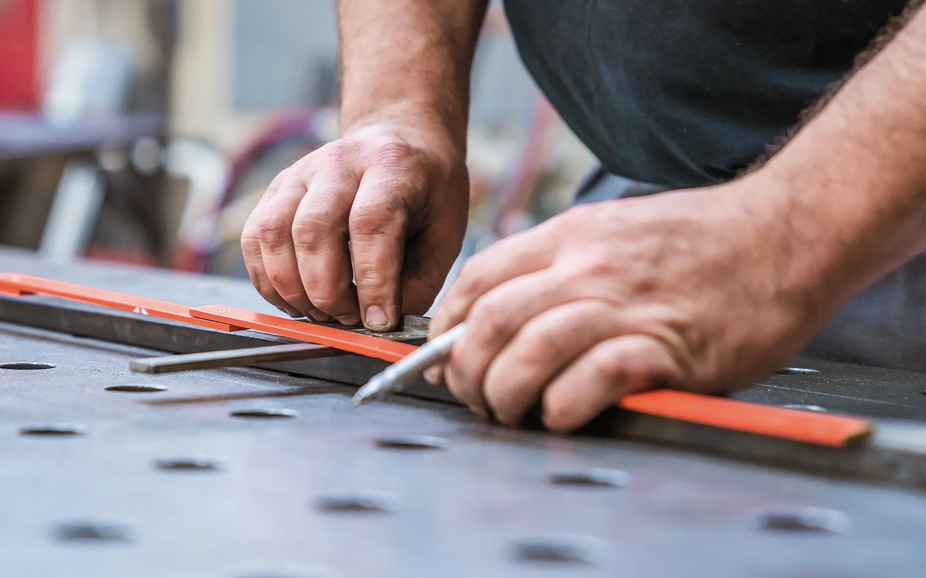 Two hands marking a steel bar against a rule on a perforated workbench